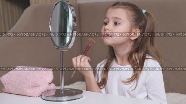 Fashion little girl in a white T-shirt is sitting at the dressing table in front of the mirror and applying makeup with face powder. Advertising of children's cosmetics concept. Footage for kids blog. Стоковое видео, видеограф Ирина Михайличенко / Фотобанк Лори