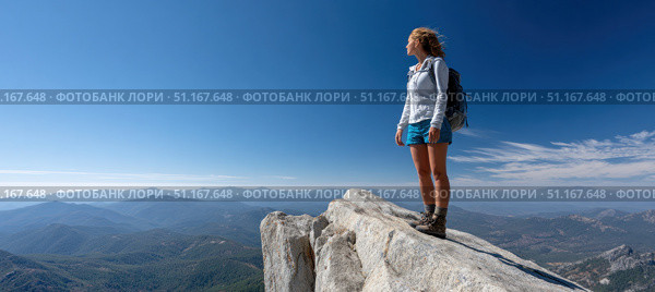 Female hiker with a backpack standing on the edge of a cliff, soaking in the breathtaking view of majestic mountain peaks under a clear blue sky on a sunny day. Стоковое AI-изображение, AI-художник Юрий Поздников / Фотобанк Лори
