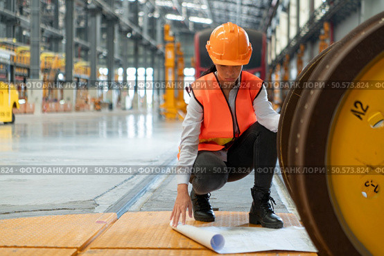 Female worker in safety vest and hardhat sitting on floor in a skytrain repair station, Using blueprints to inspect train wheels. Стоковое фото, агентство Ingram Publishing / Фотобанк Лори