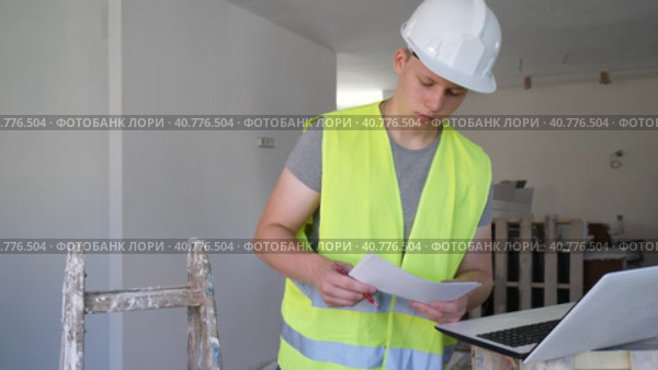 Fifteen year old worker in a yellow vest checks the house repair plan on a laptop, holding papers with drawings in his hands. Стоковое видео, видеограф Яков Филимонов / Фотобанк Лори