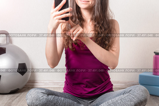 Fitness young woman sitting on the floor and using mobile phone. Fit-ball, yoga blocks and water bottle are in the background. Стоковое фото, фотограф Алдошина Екатерина / Фотобанк Лори