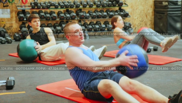 Focused adult man taking part in high-intensity group training session, doing V-sit exercise with medicine ball and twisting torso to strengthen abdominal and oblique muscles. Стоковое видео, видеограф Яков Филимонов / Фотобанк Лори