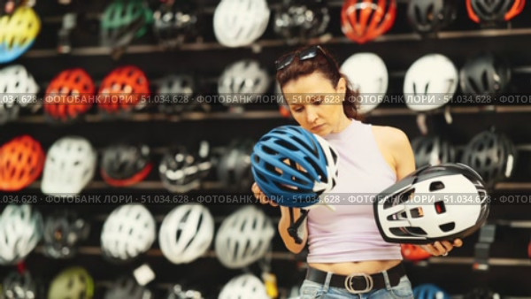 Focused interested female shopper comparing two bicycle helmets, weighing design and safety features, in well-stocked sports store with focus on cycling gear. Стоковое видео, видеограф Яков Филимонов / Фотобанк Лори
