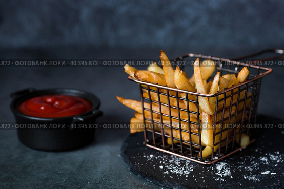 French fries in a metal basket with salt and ketchup on a stylish board on a dark background. Fried potatoes. The concept of fast food and junk food. Стоковое фото, фотограф Photojuli / Фотобанк Лори