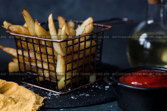 French fries in a metal basket with salt, ketchup and sunflower oil on a stylish board on a dark background. Fried potatoes. The concept of fast food and junk food. Стоковое фото, фотограф Photojuli / Фотобанк Лори