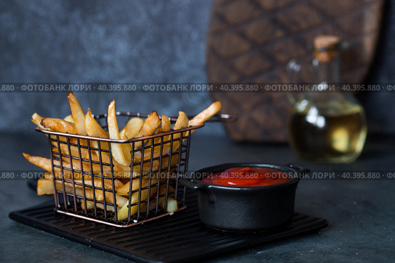 French fries in a metal basket with salt, ketchup and sunflower oil on a stylish board on a dark background. Fried potatoes. The concept of fast food and junk food. Стоковое фото, фотограф Photojuli / Фотобанк Лори