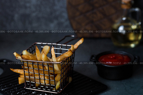 French fries in a metal basket with salt, ketchup and sunflower oil on a stylish board on a dark background. Fried potatoes. The concept of fast food and junk food. Стоковое фото, фотограф Photojuli / Фотобанк Лори
