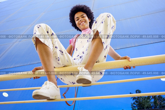 From below of delighted African American female looking at camera while sitting on railing with bent legs near glass building. Smiling black woman sitting on fence in city. Стоковое фото, агентство Ingram Publishing / Фотобанк Лори