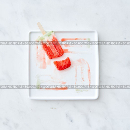 Frozen juice on a stick. Broken ice cream in a plate with splashes of ice cream on a gray marble background with copy space. Flat lay. Broken fruit ice cream lolly on a white plate with a pattern from the thawing ice cream on a gray marble background. Copy space for text. Flat lay. Стоковое фото, агентство Ingram Publishing / Фотобанк Лори