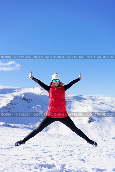 Full body of excited young female in vest beanie cap polarized sunglasses looking at camera, while leaping in air with widened legs stretched arms against snowy mountains and blue sky. Carefree young woman jumping with raised arms in snowy terrain. Стоковое фото, агентство Ingram Publishing / Фотобанк Лори