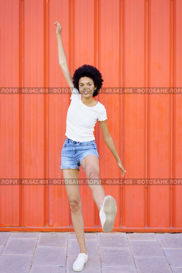 Full body of optimistic African American female in casual clothes looking at camera with raised arm while standing on one leg on red background. Glad black woman with raised leg. Стоковое фото, агентство Ingram Publishing / Фотобанк Лори