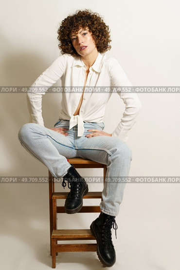Full body of self-confident woman, with curly red hair in casual outfit chilling on wooden stool and looking away against white background. Self-confident woman relaxing on ladder. Стоковое фото, агентство Ingram Publishing / Фотобанк Лори