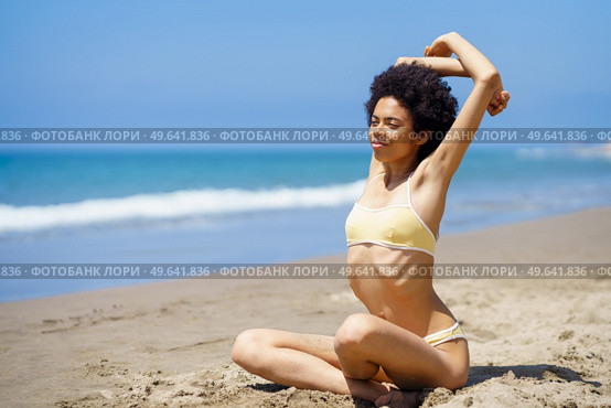 Full body of young positive African American female millennial with curly dark hair in trendy bikini, stretching arms while warming up on sandy coast against cloudless blue sky and sea. Smiling black lady stretching arms on sandy beach during summer vacation. Стоковое фото, агентство Ingram Publishing / Фотобанк Лори