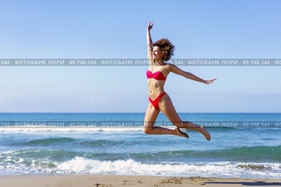 Full body side view of excited barefoot young female, in bikini with outstretched arms leaping with legs in air while looking away in daylight against rippling foamy seawater. Slim woman in bikini jumping on sandy beach. Стоковое фото, агентство Ingram Publishing / Фотобанк Лори