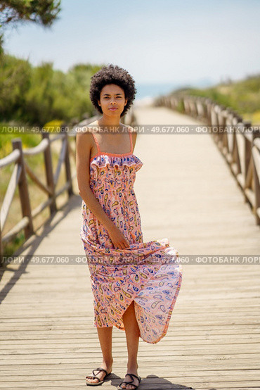 Full body young black woman with curly hair looking at camera, and waving skirt of long dress while standing on boardwalk leading to sea on sunny summer day. Ethnic tourist waving dress skirt. Стоковое фото, агентство Ingram Publishing / Фотобанк Лори