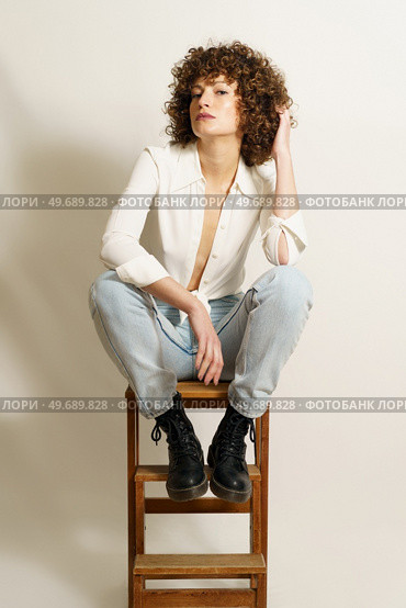 Full length of flirty woman model with curly hair, in stylish wear resting on wooden stool playing with curl and looking at camera against white background. Self confident adult female chilling on ladder. Стоковое фото, агентство Ingram Publishing / Фотобанк Лори