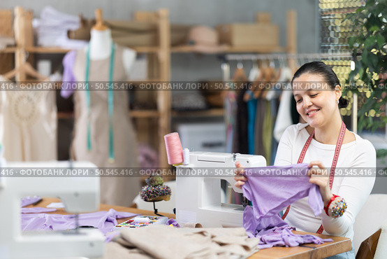 Glad young Asian tailoress viewing dress detail sitting at sewing-machine. Стоковое фото, фотограф Яков Филимонов / Фотобанк Лори