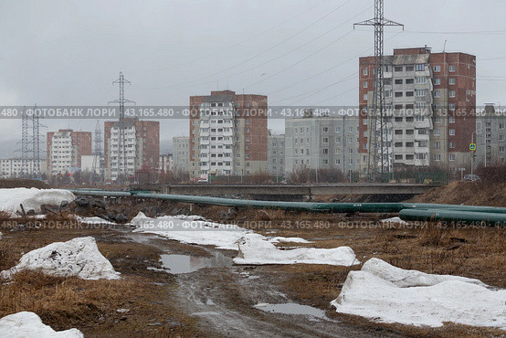 Город Магадан, Магаданская область, Россия. Вид на многоэтажные жилые дома на окраине города. На переднем плане грунтовая дорога, трубопровод, лужи и тающие сугробы снега. Дождливая весенняя погода. Май. (2024 год). Редакционное фото, фотограф Андрей Степанов / Фотобанк Лори