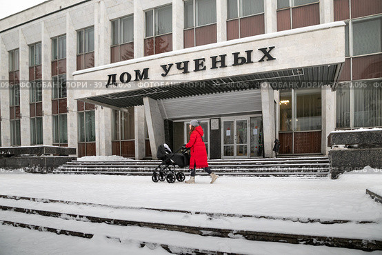 Город Обнинск, проспект Ленина, 129. Девушка в красном пальто с детской коляской идет мимо Дома ученых (МП "Дом ученых") в зимний пасмурный день (2021 год). Редакционное фото, фотограф Наталья Николаева / Фотобанк Лори