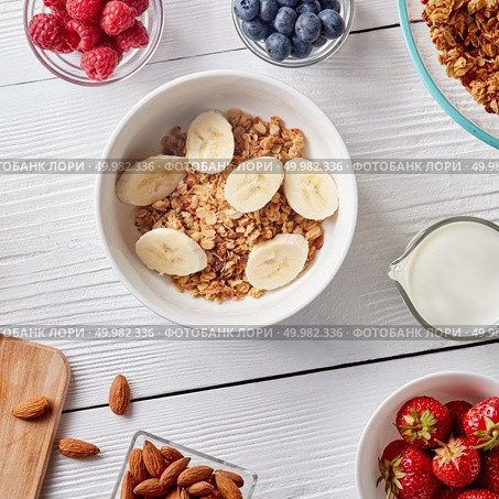 Grain free oat free paleo granola mixed with bananas slice, in bowl with nuts, raspberries, strawberries, blueberries and milk in a decanter on a white wooden background. Healthy breakfast ingredients on a white woodwn background. Granola, nuts, berries, milk, banana in bowl , top view. Стоковое фото, агентство Ingram Publishing / Фотобанк Лори