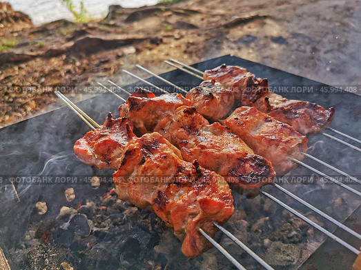 Grilling skewered meat over an open flame at a picturesque riverside during a sunny afternoon. Стоковое фото, фотограф Александра Селина / Фотобанк Лори