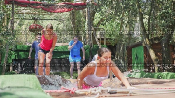 Group of cheerful young adults having fun in green summer amusement park, moving through obstacle course with soft pillows in pool of water. Стоковое видео, видеограф Яков Филимонов / Фотобанк Лори
