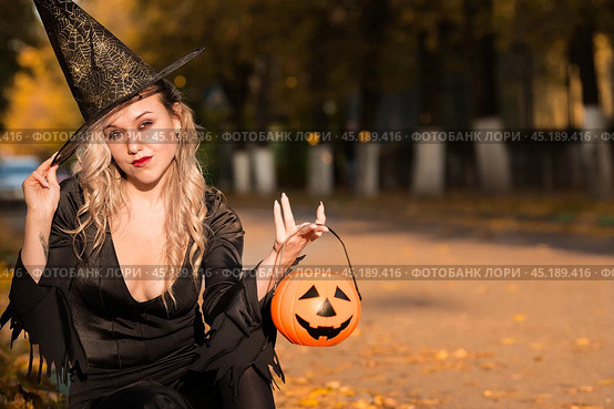 Halloween Witch with pumpkin. Beautiful young woman in a witch costume on the street. Halloween decor. Copy space. Стоковое фото, фотограф Photojuli / Фотобанк Лори