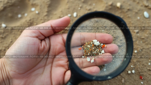 Hand holding a magnifying glass over a pile of sand. Microplastic. Стоковое AI-изображение, AI-художник Sergey Grishin / Фотобанк Лори