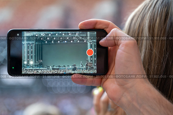 Hand of unidentified man records video with mobile smart phone of concert stage. Crowd of fans with smartphone in hands enjoy live music at concert in vivid stage lights at evening. Стоковое фото, фотограф Данила Большаков / Фотобанк Лори