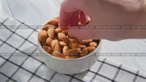 Hand picking an almond from a white ceramic bowl overflowing with almonds, resting on a checkered tablecloth atop a light wooden table, highlighting healthy snacking options. Стоковое видео, видеограф Ольга Ильина / Фотобанк Лори