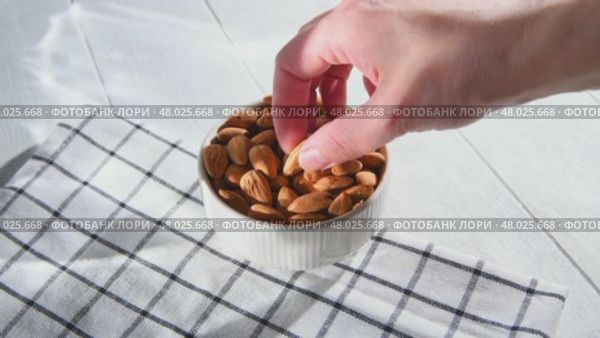 Hand picking an almond from a white ceramic bowl overflowing with almonds, resting on a checkered tablecloth atop a light wooden table, highlighting healthy snacking options. Стоковое видео, видеограф Ольга Ильина / Фотобанк Лори