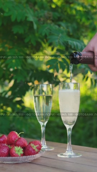 Hand pours sparkling wine into flutes on wooden table, next to bowl of fresh strawberries with lush green background. Ideal for themes of celebration, summer and romance. Стоковое видео, видеограф Елена Никишина / Фотобанк Лори