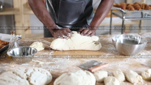 Hands of professional African baker in maroon apron kneading dough on floury wooden table, filled with baking tools and ingredients, in artisan bakery shop. Стоковое видео, видеограф Яков Филимонов / Фотобанк Лори