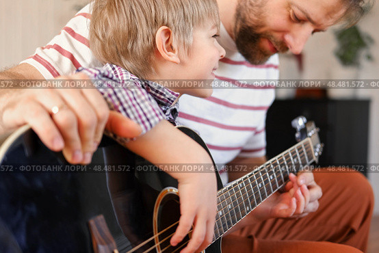 Handsome dad teaches his cute son to play the guitar sitting on sofa in the living room at home. Happy Father's Day. Father teaching his child to play guitar. Kid Learning music skill. Art education. Стоковое фото, фотограф Марина Демидюк / Фотобанк Лори