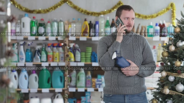 Handsome man speaking mobile and choose something in cosmetics store decorated for New Year holiday. Стоковое видео, видеограф Яков Филимонов / Фотобанк Лори