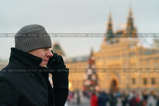 Handsome man walking in the Moscow city center during coronavirus pandemic. New Year holidays. He talking on the smartphone. The conversation is in a positive mood. GUM (Central department store) (2021 год). Стоковое фото, фотограф YuryKara / Фотобанк Лори