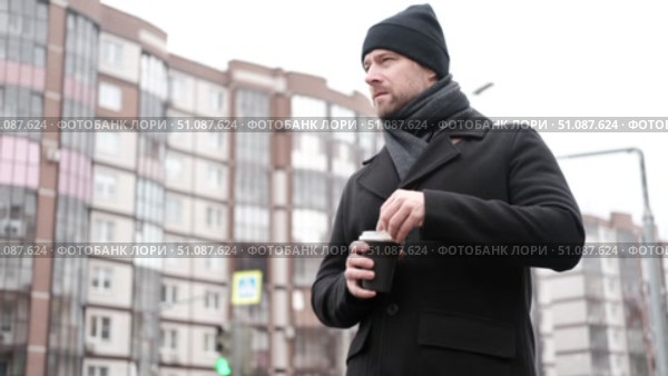 Handsome man wearing black coat and brown leather bag walking down on the city, drinking coffee paper takeaway cup with mobile phone. Stylish bearded middle-aged male using smartphone at urban street. Стоковое видео, видеограф Марина Демидюк / Фотобанк Лори