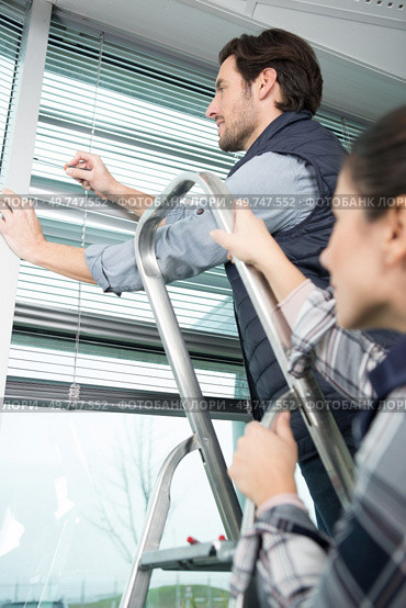 handyman repairing window blind his partner holding the stepladder. Стоковое фото, агентство Ingram Publishing / Фотобанк Лори