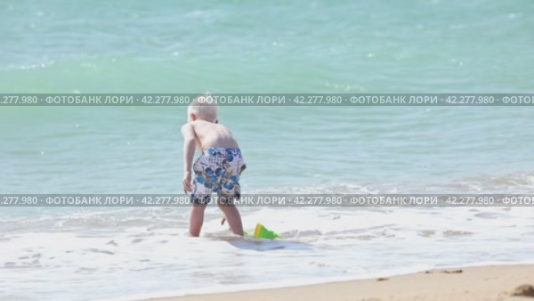 Happy child boy playing with the sand on the beach, pouring water from plastic bucket on the sea beach, building sand castle, enjoying summer. Advertising for tour operators, hotels, airlines. Стоковое видео, видеограф Ирина Михайличенко / Фотобанк Лори