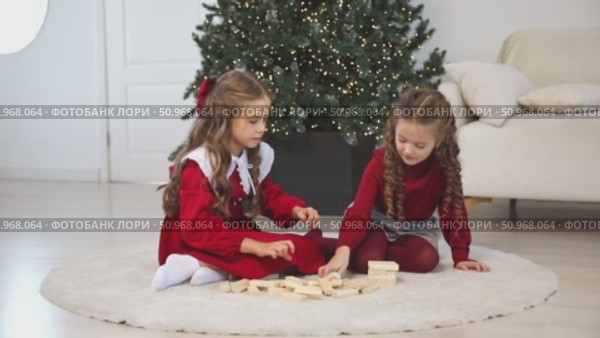 Happy family, little sister girls or best friends are sitting on floor near Xmas tree, playing board game with wooden tower together at home while spending leisure Christmas holidays. Footage. Стоковое видео, видеограф Ирина Михайличенко / Фотобанк Лори
