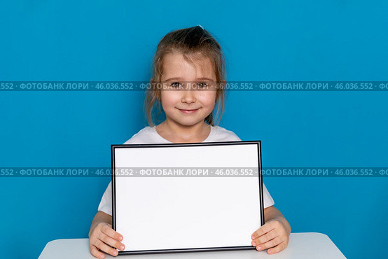Happy little preschool girl sitting at table with empty white mock up paper board on a light blue background, copy space. Preschool education, goods for children. High quality photo. Стоковое фото, фотограф Ирина Михайличенко / Фотобанк Лори