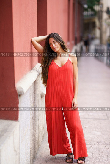 In a vibrant urban setting, a stylish woman in a flowing red jumpsuit poses elegantly, exuding charm and sophistication, attracting the admiration of curious onlookers who admire her fashion sense. An Elegant Woman Dressed in a Stunning Red Jumpsuit Posing Gracefully Against a City Wall. Стоковое фото, агентство Ingram Publishing / Фотобанк Лори