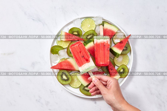 In the female hand, a healthy berry icy popsicle against the background of a gray marble table with a plate with slices of watermelon, kiwi, lime and ice cubes. Space for text. Flat lay. A woman’s hand holds a frozen watermelon smoothie on a stick against the background of a plate with pieces of fruit and ice cubes on a gray marble table. Copy space. Flat lay. Стоковое фото, агентство Ingram Publishing / Фотобанк Лори