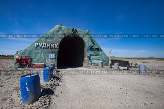 Khromtau, Aktobe region, Kazakhstan - May 06 2012: Gold ore mine portal. Title: Quarry and Mechel company logo. Blue plastic barrel with concrete. Blue sky and desert road. Редакционное фото, фотограф Alexey Rezvykh / Фотобанк Лори