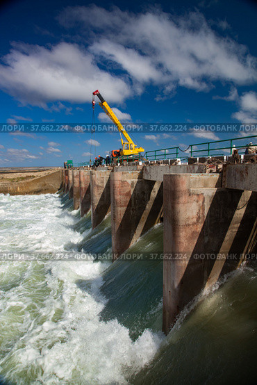 Kok-aral, Kazakhstan - May 03, 2012: Small Aral Sea Kok-aral dam. Water discharge in flood. Open shutters. Yellow mobile crane and workers on dam. Стоковое фото, фотограф Alexey Rezvykh / Фотобанк Лори