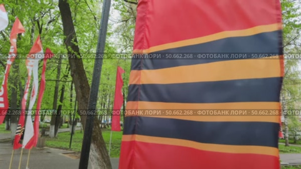 Красный флаг с Георгиевской лентой развивается на ветру в городском парке. Праздничное оформление города ко Дню Победы в Великой Отечественной Войне 9 мая. Red flag with St. George ribbon is fluttering in wind. Festive decoration of city for Victory Day in Great Patriotic War on May 9. Inscriptions on flags in Russian are word Victory. Стоковое видео, видеограф Ирина / Фотобанк Лори