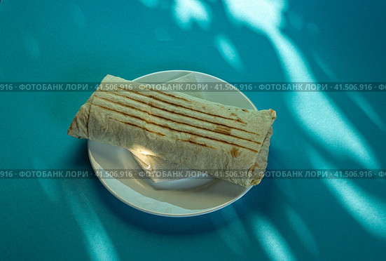 Large shawarma on a white plate with a napkin on a blue background with shadows. Food, healthy fast food, delicious to eat. Стоковое фото, фотограф Tae Mi / Фотобанк Лори