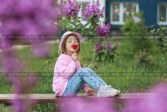 Lifestyle. A beautiful little girl in pink clothes with a lollipop in the shape of a heart. The girl licks the candy. The concept of love and sweets. Стоковое фото, фотограф Photojuli / Фотобанк Лори