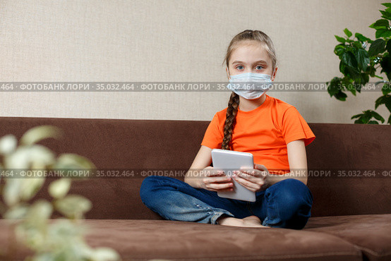 Little girl in medical mask sitting on the sofa in the Lotus position and using tablet pc. Isolation at home. Looking into the camera. Стоковое фото, фотограф Иван Карпов / Фотобанк Лори
