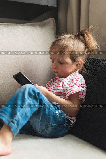 Little girl is sitting on sofa with smart phone. She's watching intently in it. Стоковое фото, фотограф Алдошина Екатерина / Фотобанк Лори